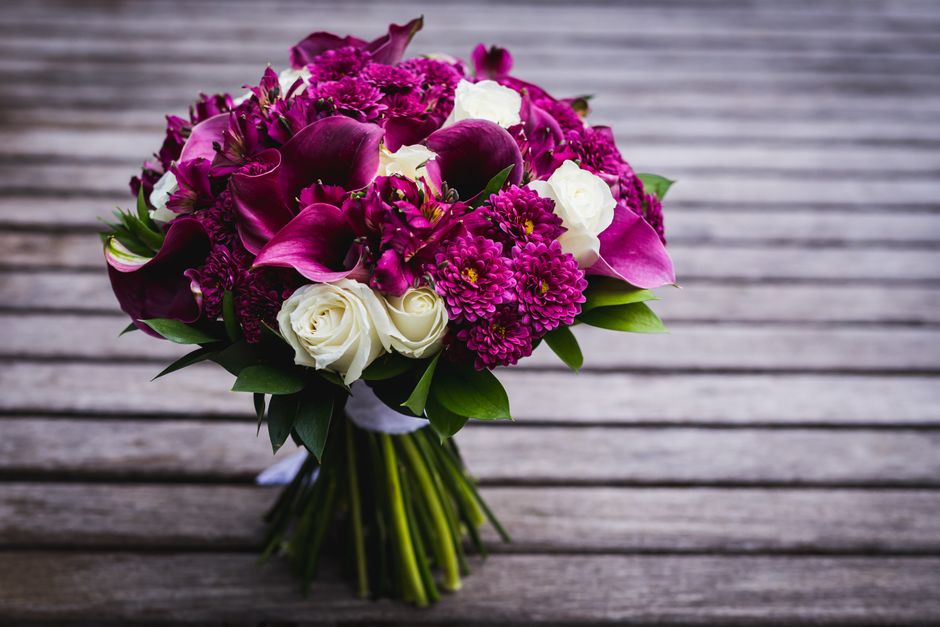 A vibrant bouquet of fresh flowers arranged with deep pink calla lilies, purple chrysanthemums, and white roses, accented with green leaves. The flowers are neatly packed in a rounded style and wrappe