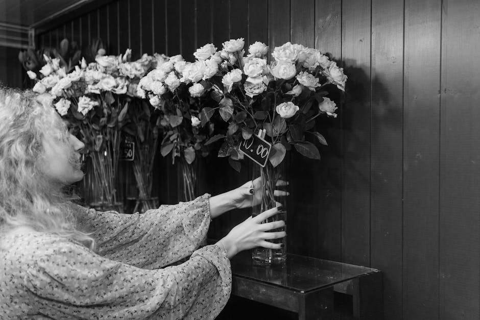 A woman with curly hair wearing a patterned long-sleeve blouse is adjusting a large bouquet of fresh white roses and greenery in a tall glass vase, positioned on a small wooden table against a dark wo
